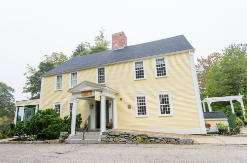 A yellow two-story house with white trim, a front porch, and a stone pathway, surrounded by greenery.