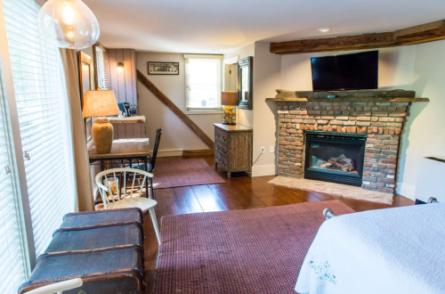 Cozy living space with a brick fireplace, wooden furniture, and a desk, featuring natural light from windows.
