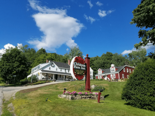 A scenic view of the Christmas Farm Inn with a sign, surrounded by green grass and trees under a blue sky.
