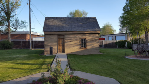 A small, rustic building with a sloped roof, surrounded by green grass and trees, with a pathway leading to it.