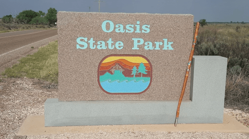 Sign for Oasis State Park featuring a colorful logo and a walking stick leaning against it, set against a rural backdrop.