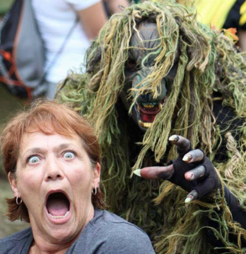 A surprised woman gasps as a person in a creepy, nature-themed costume looms behind her, both expressing strong emotions.