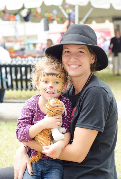 A woman in a black hat smiles while holding a girl with face paint as a tiger, both sitting outdoors.
