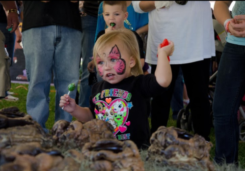 A young child with face paint and a lollipop stands among a crowd, holding a toy while looking excited.