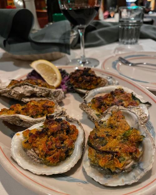 A plate of baked oysters topped with a golden crust, garnished with lemon and served with a glass of red wine.
