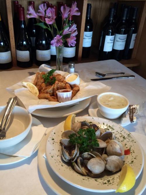 A table setting with fried seafood, clam chowder, and a vase of flowers, surrounded by bottles of wine in the background.