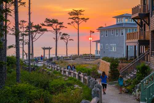 A scenic sunset over the ocean, with people walking along a path beside beach houses and trees.