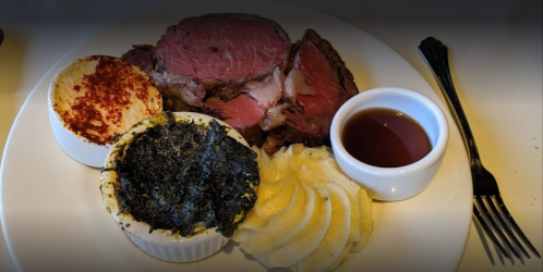 A plate featuring sliced prime rib, creamy mashed potatoes, spinach dip, and a small cup of sauce.