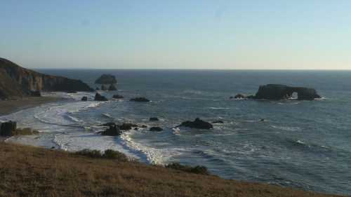 A serene coastal view with waves crashing on rocky shores and distant sea stacks under a clear blue sky.