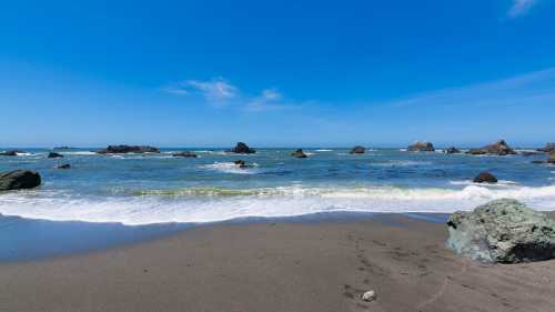 A serene beach scene with gentle waves, rocky outcrops, and a clear blue sky.