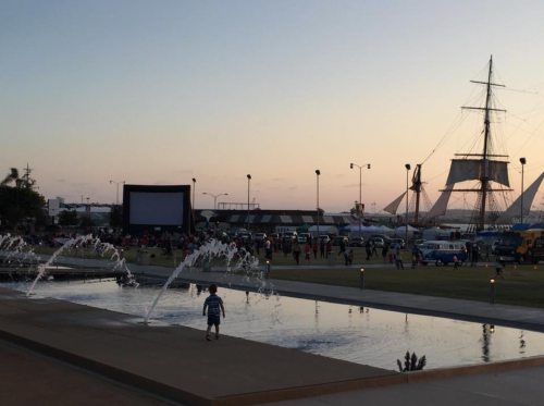 A sunset view of a waterfront park with a large screen, fountain, and a tall ship in the background.