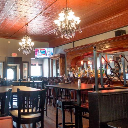 Interior of a restaurant featuring wooden tables, chandeliers, a bar, and a TV displaying a sports event.