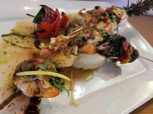 Grilled shrimp skewers with vegetables, drizzled in sauce, served on a white plate with herbs and lemon slices.