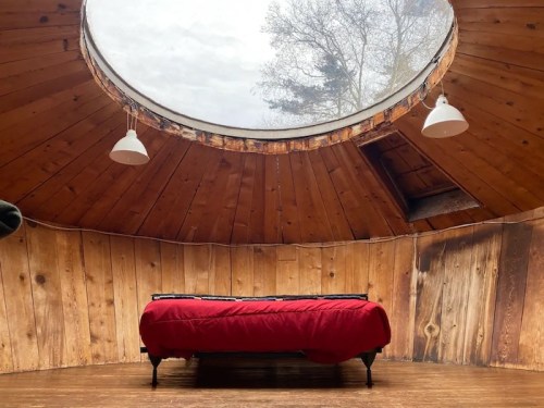 A cozy circular room with wooden walls, featuring a bed covered in a red blanket and a skylight above.