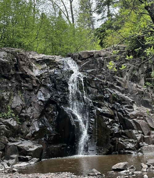 A small waterfall cascades over rocky terrain into a calm pool, surrounded by lush green trees.