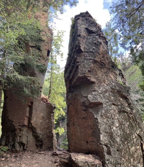 Two tall, rugged rock formations surrounded by lush green trees and a bright blue sky.