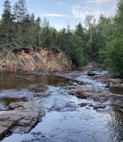 A serene river flows past rocky cliffs, surrounded by lush green trees under a clear blue sky.