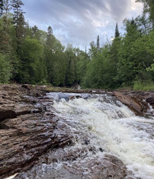 A rushing river flows over rocky terrain, surrounded by lush green trees and a cloudy sky in the background.