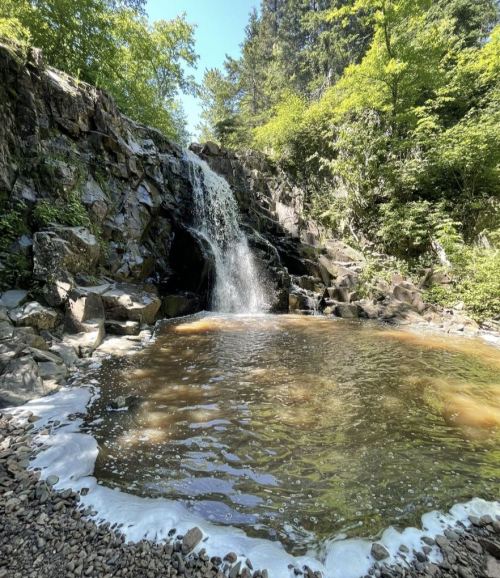 A serene waterfall cascades over rocky terrain into a calm, brown pool surrounded by lush greenery.