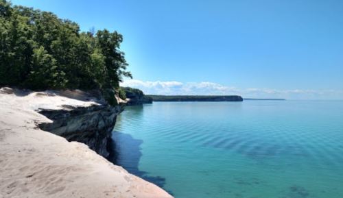 A serene shoreline with sandy beach, clear blue water, and lush green trees under a bright blue sky.