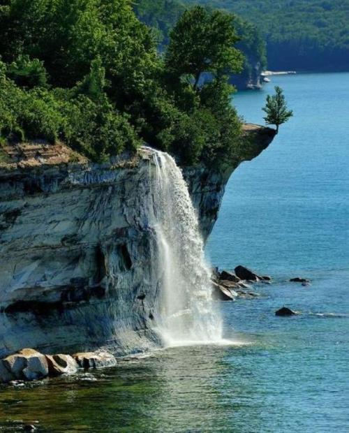 A waterfall cascades over a rocky cliff into a serene blue lake, surrounded by lush green trees.