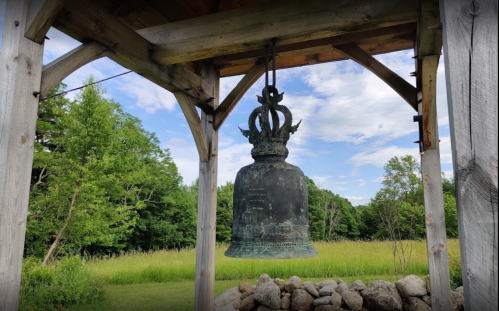 A large, ornate bell hangs from a wooden structure, surrounded by green trees and a grassy field under a blue sky.