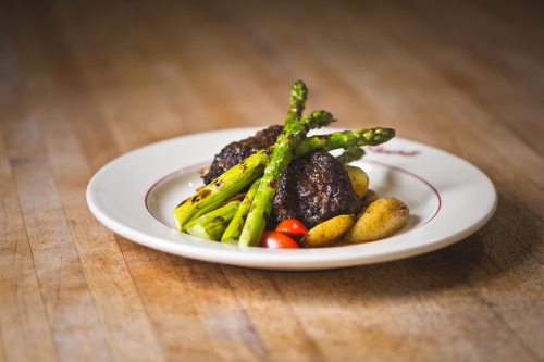 A plate of grilled meat patties served with asparagus, cherry tomatoes, and roasted potatoes on a wooden table.