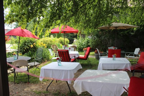 Outdoor dining area with white tables, red chairs, and umbrellas, surrounded by greenery and flowers.