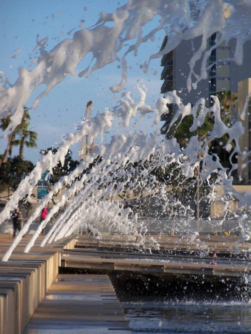 A fountain with water jets creating splashes, set against a backdrop of buildings and palm trees.