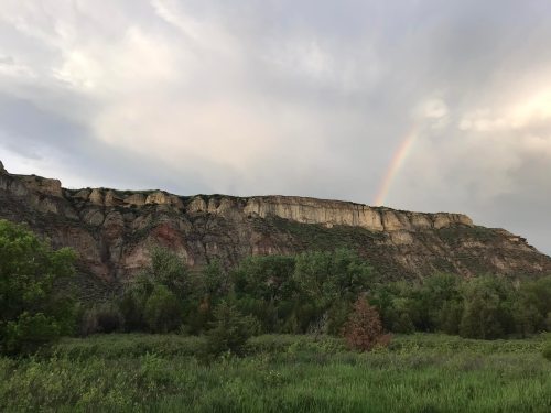 A scenic view of a cliff with greenery in the foreground and a rainbow arching across a cloudy sky.