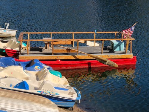 A red boat with a wooden frame and an oar, docked beside several colorful paddle boats on calm water.