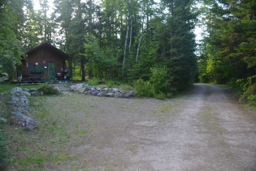 A rustic cabin surrounded by trees, with a gravel path leading into the woods.