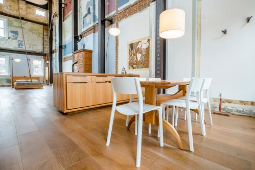A modern dining area with a wooden table, white chairs, and large windows, featuring exposed brick and artwork on the walls.