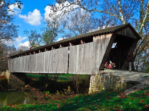 A wooden covered bridge spans a serene river, surrounded by trees and a clear blue sky.