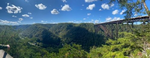 A panoramic view of a lush green valley with a large bridge spanning across it under a blue sky with fluffy clouds.