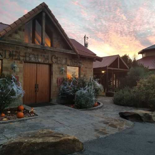 A rustic winery building with pumpkins and spiderweb decorations, set against a colorful sunset sky.