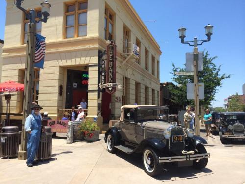 A vintage car parked in front of a historic bank building, with people and street lamps in a sunny setting.