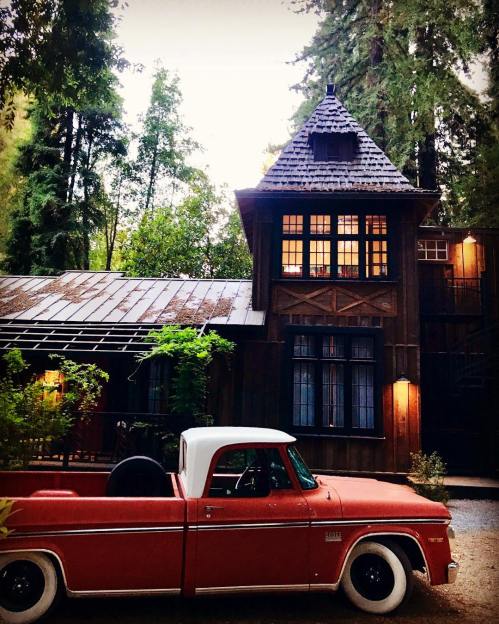 A vintage red pickup truck parked in front of a charming wooden house surrounded by tall trees.