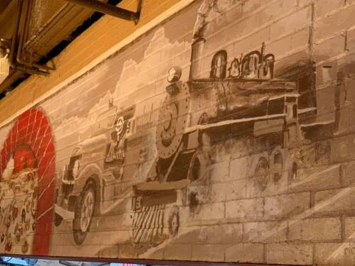 A mural depicting a vintage train and an old car on a brick wall, with a cloudy sky in the background.
