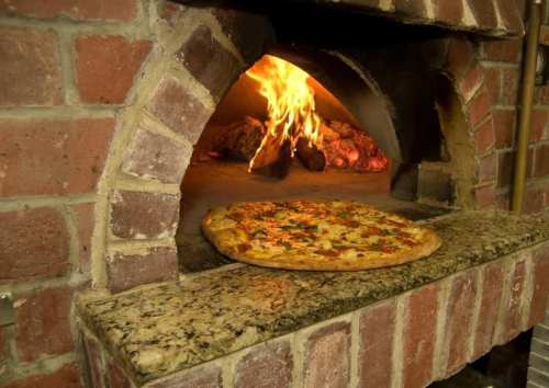A freshly baked pizza sits on a stone ledge in front of a brick oven with flames visible inside.