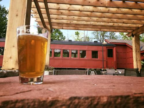A glass of beer sits on a stone ledge, with a red train car and wooden pergola in the background.