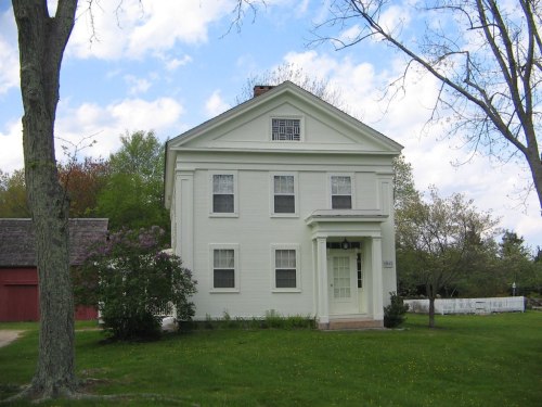 A white two-story house with a gabled roof, surrounded by trees and a grassy area under a partly cloudy sky.