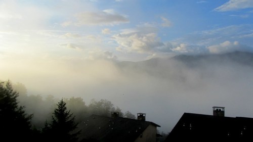 A misty landscape with low clouds, mountains in the background, and rooftops partially obscured by fog.