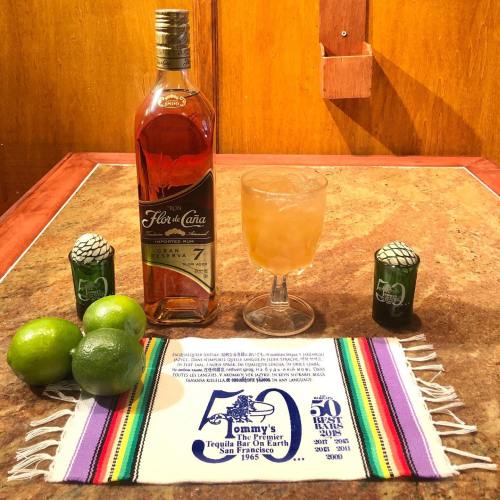 A bottle of Flor de Caña rum, a cocktail, limes, and decorative items on a colorful table runner celebrating Tommy's Tequila Bar.