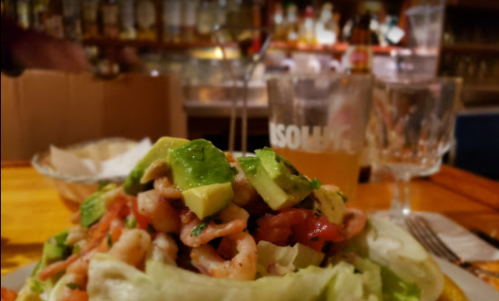A colorful shrimp salad with avocado on a bed of lettuce, set against a blurred bar background.
