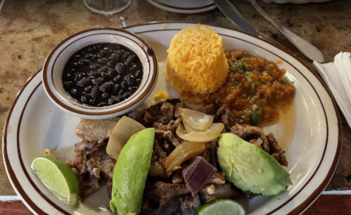 A plate of grilled meat with onions, avocado, lime, rice, black beans, and a vegetable medley.