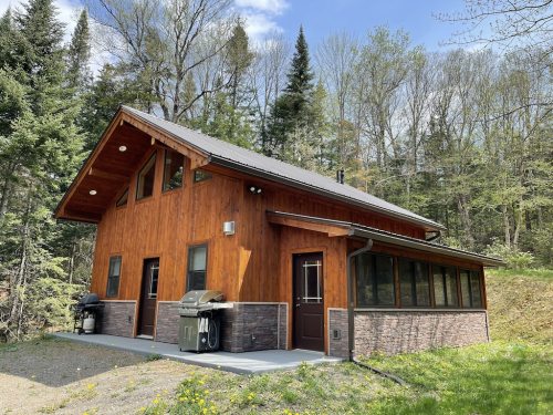 A modern wooden cabin surrounded by trees, featuring a stone foundation and a patio with outdoor grills.