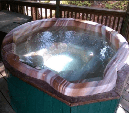 A wooden hot tub filled with clear water, surrounded by a wooden deck and greenery in the background.