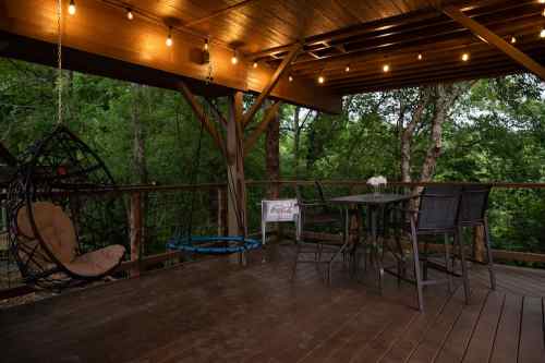 A cozy outdoor deck with a hanging chair, table, and chairs, surrounded by trees and string lights.