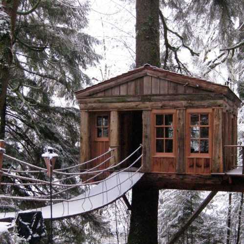 A rustic treehouse nestled among tall trees, connected by a wooden bridge, surrounded by snow-covered ground.